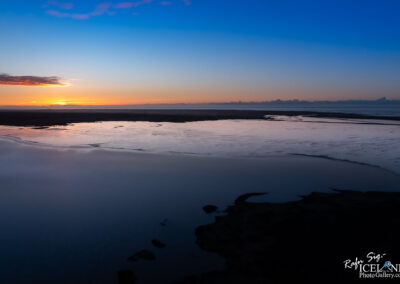 The image depicts a serene coastal landscape during sunset. The sky transitions from vibrant oranges and yellows near the horizon to deep blues higher up. The water reflects these colors, creating a beautiful, glimmering surface. In the foreground, there are patches of land visible, bordered by calm water, which likely represents tidal flats or a calm inlet. The atmosphere feels peaceful and tranquil, evoking a sense of natural beauty and stillness.