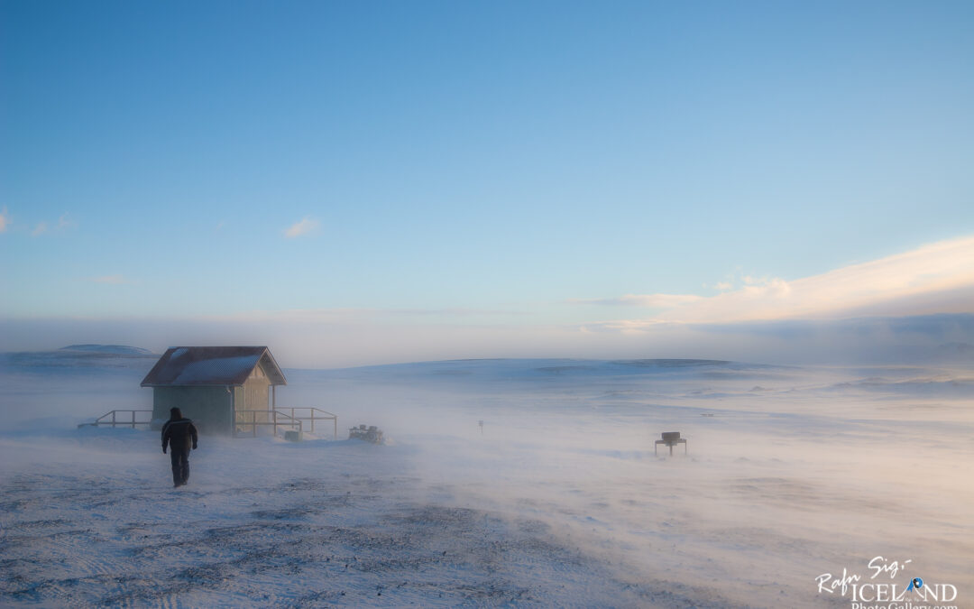 A lone figure walks toward a small, green, wooden cabin with a sloped roof, set against a vast, snowy landscape. The ground is covered in a thick layer of snow, and a light mist rises as the wind sweeps across the scene. In the background, rolling hills partially emerge from the frost, under a bright blue sky streaked with soft clouds. The atmosphere conveys a sense of solitude and harsh beauty, highlighting the stark contrast between the cabin and the surrounding winter wilderness.