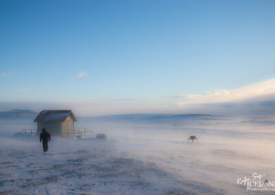 A lone figure walks toward a small, green, wooden cabin with a sloped roof, set against a vast, snowy landscape. The ground is covered in a thick layer of snow, and a light mist rises as the wind sweeps across the scene. In the background, rolling hills partially emerge from the frost, under a bright blue sky streaked with soft clouds. The atmosphere conveys a sense of solitude and harsh beauty, highlighting the stark contrast between the cabin and the surrounding winter wilderness.