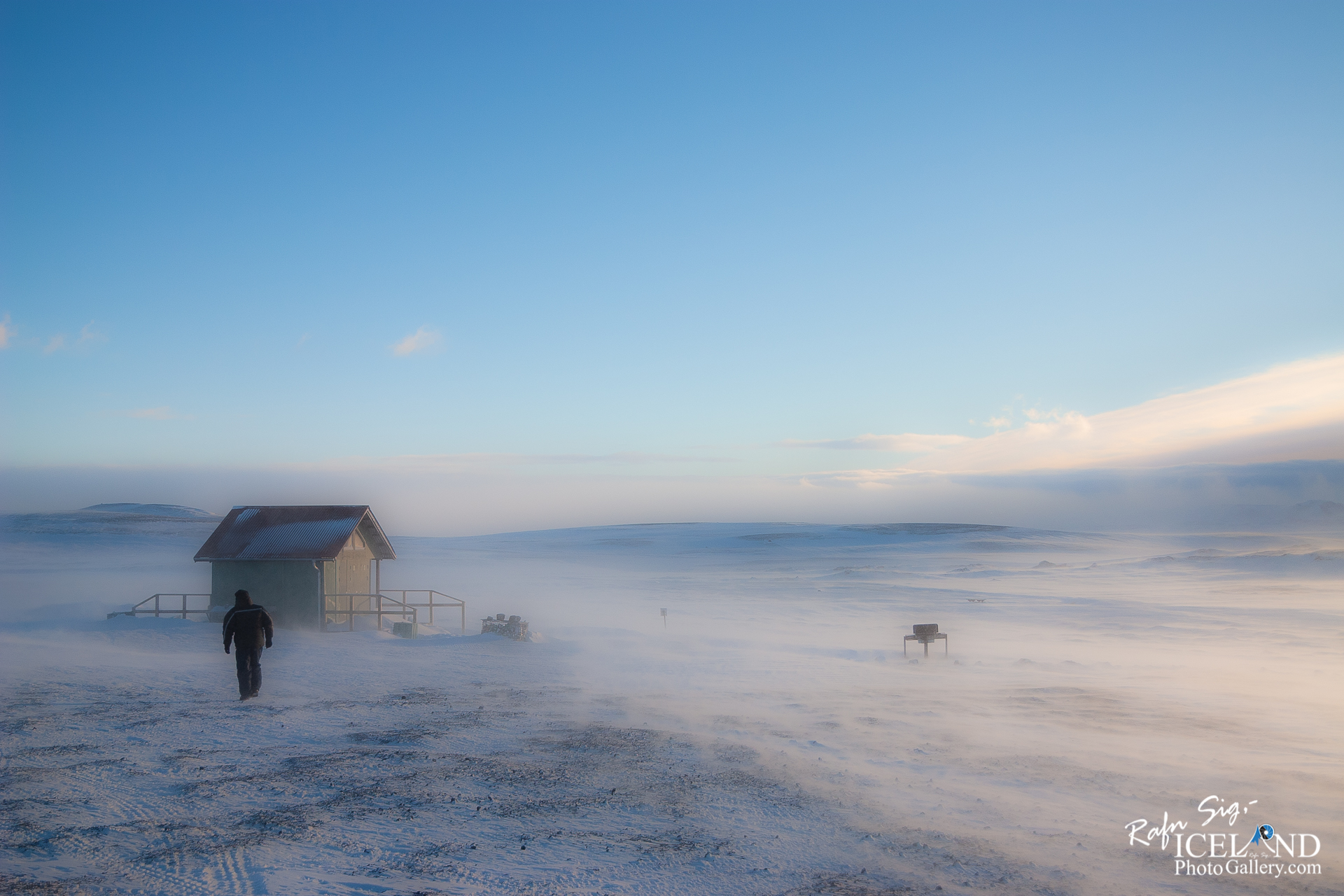A lone figure walks toward a small, green, wooden cabin with a sloped roof, set against a vast, snowy landscape. The ground is covered in a thick layer of snow, and a light mist rises as the wind sweeps across the scene. In the background, rolling hills partially emerge from the frost, under a bright blue sky streaked with soft clouds. The atmosphere conveys a sense of solitude and harsh beauty, highlighting the stark contrast between the cabin and the surrounding winter wilderness.