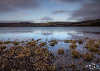 A serene lakeside scene featuring calm water that reflects the sky above, creating a mirror-like effect. The foreground has patches of grass and rocky terrain edging the water. In the background, gentle hills rise, partially covered in soft green vegetation and fading into the horizon. The sky is a mix of light blue and scattered clouds, suggesting a peaceful, early morning or late afternoon atmosphere. The overall mood conveys tranquility and natural beauty.