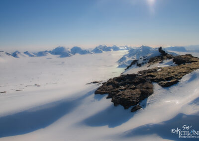 A breathtaking winter landscape stretches out, featuring a vast expanse of snow-covered plains that glisten in bright sunlight. In the background, majestic mountains rise with their peaks capped in snow, creating a dramatic contrast against a clear blue sky. The gentle slopes and ridges of the terrain exhibit subtle shadows, adding depth to the scene. A rocky foreground juts out towards the viewer, showcasing dark, rugged stones that are partially covered in patches of moss, suggesting a rich ecosystem beneath the snow. The overall atmosphere conveys a sense of serenity and awe in nature's grandeur.