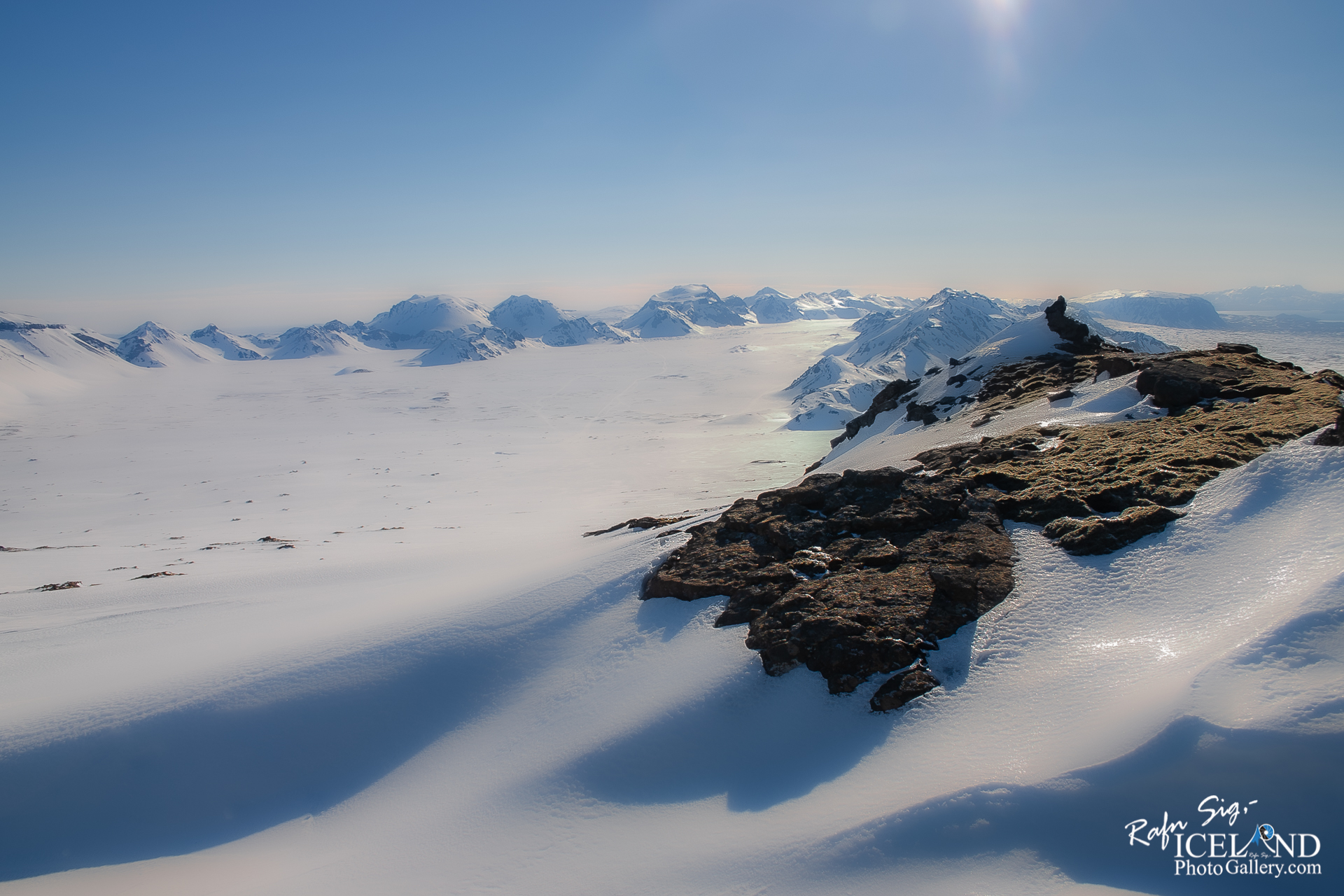 A breathtaking winter landscape stretches out, featuring a vast expanse of snow-covered plains that glisten in bright sunlight. In the background, majestic mountains rise with their peaks capped in snow, creating a dramatic contrast against a clear blue sky. The gentle slopes and ridges of the terrain exhibit subtle shadows, adding depth to the scene. A rocky foreground juts out towards the viewer, showcasing dark, rugged stones that are partially covered in patches of moss, suggesting a rich ecosystem beneath the snow. The overall atmosphere conveys a sense of serenity and awe in nature's grandeur.