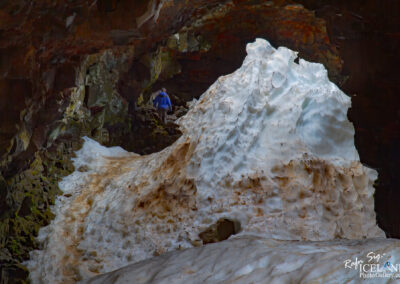 A person in a blue jacket climbs a rocky path leading into a dark cave. The path is lined with patches of ice and snow that appear dirty and textured, contrasting with the dark, rugged walls of the cave. The rocks have a mixture of green moss and earthy tones, highlighting the natural environment. In the background, the cave opens up further, hinting at more rocky formations. The overall scene conveys a sense of adventure in a remote, icy landscape.