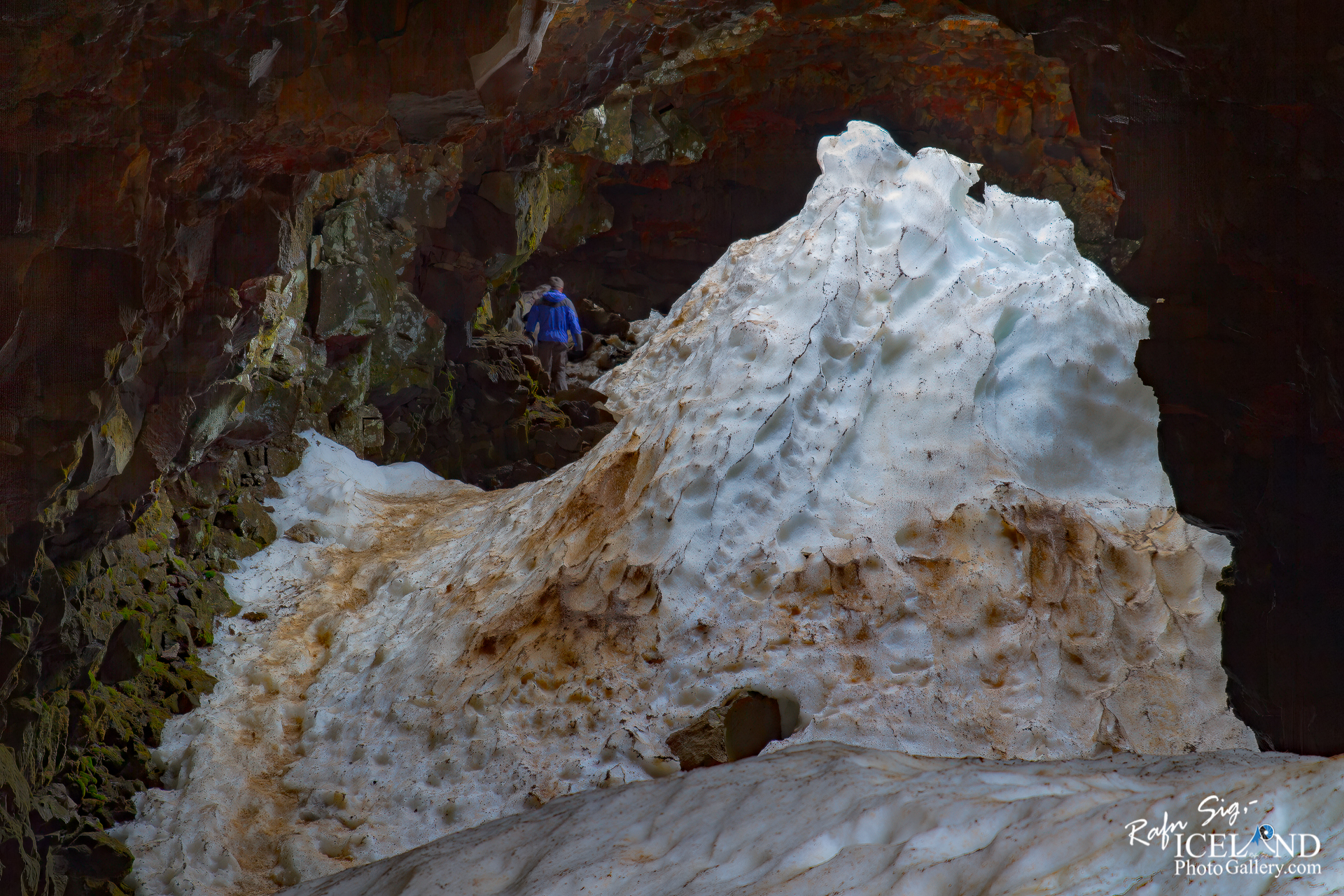 A person in a blue jacket climbs a rocky path leading into a dark cave. The path is lined with patches of ice and snow that appear dirty and textured, contrasting with the dark, rugged walls of the cave. The rocks have a mixture of green moss and earthy tones, highlighting the natural environment. In the background, the cave opens up further, hinting at more rocky formations. The overall scene conveys a sense of adventure in a remote, icy landscape.