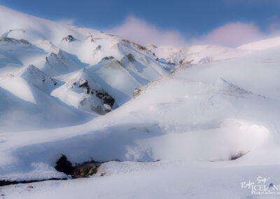 A vast, snowy landscape stretches across the scene, with gently rolling white hills and steep, jagged peaks emerging in the background. The snow glistens under a soft, blue sky, punctuated by a few wispy clouds. In the foreground, a dark crevice reveals rocky textures and streams, contrasting with the bright snow. The overall ambiance evokes a serene, wintry wilderness, showcasing the beauty of untouched nature.