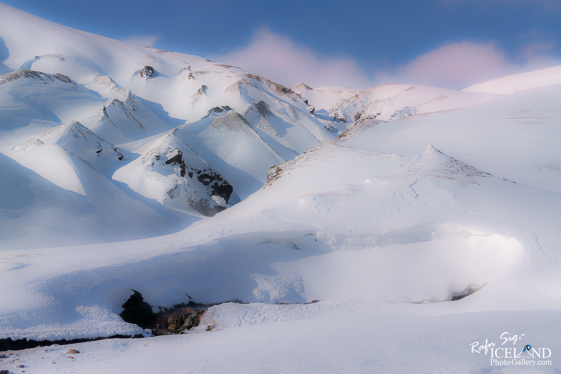 A vast, snowy landscape stretches across the scene, with gently rolling white hills and steep, jagged peaks emerging in the background. The snow glistens under a soft, blue sky, punctuated by a few wispy clouds. In the foreground, a dark crevice reveals rocky textures and streams, contrasting with the bright snow. The overall ambiance evokes a serene, wintry wilderness, showcasing the beauty of untouched nature.