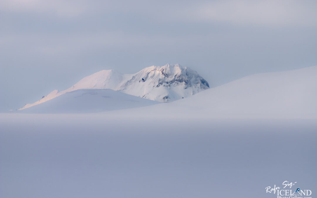A serene, snowy landscape dominates the scene, with smooth, undulating hills blanketed in white snow. In the background, a distant mountain peak is visible, also coated in snow but with jagged edges that break the smoothness of the hills in the foreground. The sky above is soft and overcast, with muted shades of light gray and hints of pastel colors, suggesting a calm and cool atmosphere. The overall impression is one of tranquility and vastness, with the snow-covered terrain extending into the distance where the mountain rises.