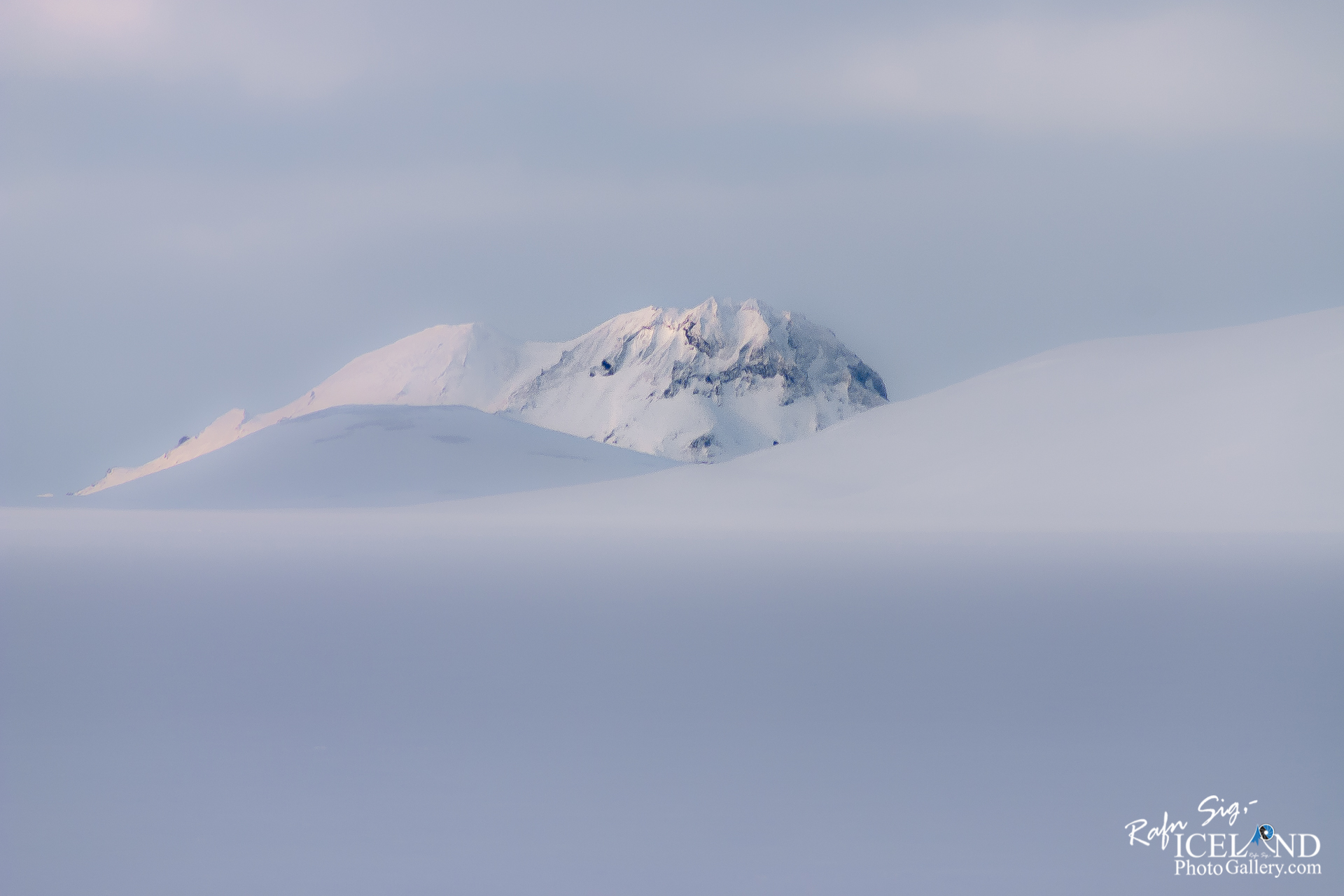 A serene, snowy landscape dominates the scene, with smooth, undulating hills blanketed in white snow. In the background, a distant mountain peak is visible, also coated in snow but with jagged edges that break the smoothness of the hills in the foreground. The sky above is soft and overcast, with muted shades of light gray and hints of pastel colors, suggesting a calm and cool atmosphere. The overall impression is one of tranquility and vastness, with the snow-covered terrain extending into the distance where the mountain rises.