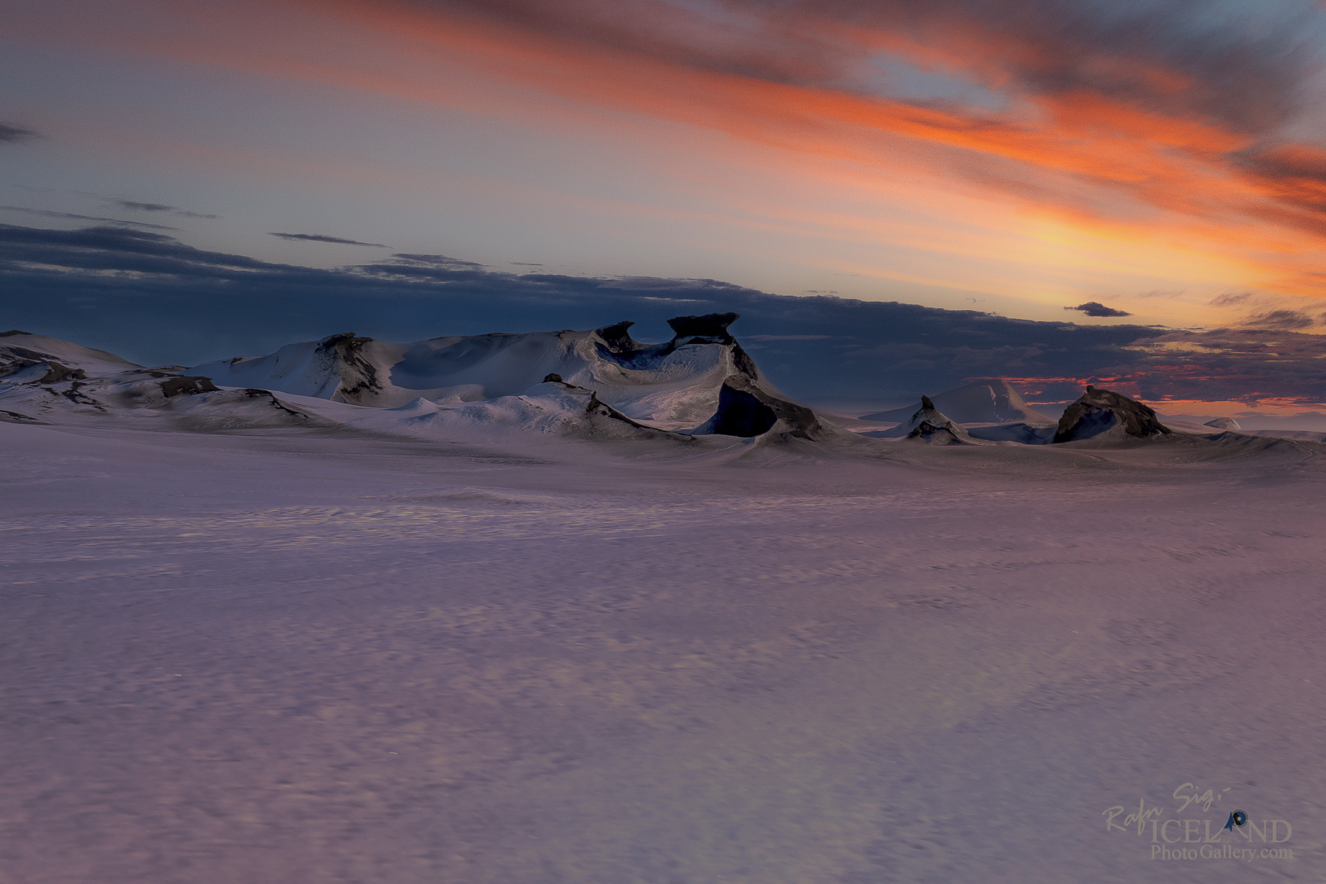 A serene lakeside scene featuring calm water that reflects the sky above, creating a mirror-like effect. The foreground has patches of grass and rocky terrain edging the water. In the background, gentle hills rise, partially covered in soft green vegetation and fading into the horizon. The sky is a mix of light blue and scattered clouds, suggesting a peaceful, early morning or late afternoon atmosphere. The overall mood conveys tranquility and natural beauty.