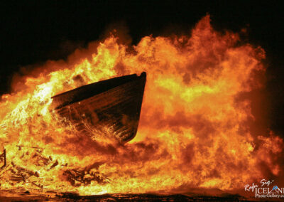 A large boat is engulfed in vibrant flames, creating a dramatic contrast against the dark night sky. The boat, positioned at the center, has a rounded bow sharply cutting through the fire, while the flames billow up and outward, casting a warm glow. The fiery orange and yellow hues of the flames blend with darker shades, giving a sense of intensity and chaos. Surrounding debris, possibly remnants of wood, is illuminated by the fire, contributing to the overall sense of destruction and drama in the scene.