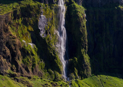 A stunning natural scene featuring a tall waterfall cascading down a rocky cliff, surrounded by lush green grass and moss. The waterfall emerges from a steep cliff face, creating a sense of tranquility as the water flows smoothly down. Below the cliff, the landscape includes patches of rocks and an expansive grassy field, punctuated by a bright red tractor working in the foreground. The sky above is a mix of blues with fluffy white clouds, enhancing the serene and vibrant atmosphere of the setting.