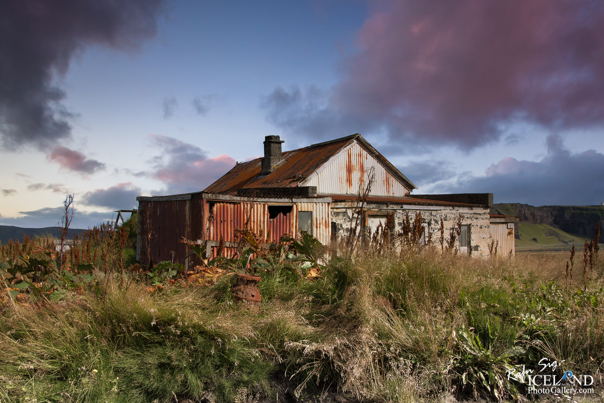 The image depicts a weathered, abandoned house surrounded by tall, overgrown grass and shrubbery. The structure features a mix of rusty red and white corrugated metal siding, with peeling paint and signs of age. Its roof, partially covered in rust, includes a chimney. In the background, a rocky hillside is visible, under a sky filled with soft, colorful clouds transitioning from pink to blue as the sun sets. The area conveys a sense of decay and solitude, with nature gradually reclaiming the space around the house.