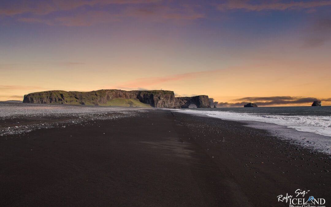 A dramatic coastal scene featuring a wide expanse of black sand beach. In the distance, a rugged cliff rises, partly covered in greenery, with a large rock formation that has a natural arch. The sky above displays a gradient of soft colors, transitioning from warm peach to cool blue as the sun sets. Gentle waves lap at the shore, and scattered pebbles dot the beach, enhancing the serene and untouched feeling of the landscape.