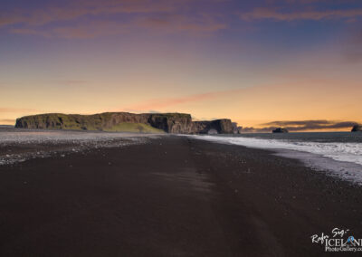 A dramatic coastal scene featuring a wide expanse of black sand beach. In the distance, a rugged cliff rises, partly covered in greenery, with a large rock formation that has a natural arch. The sky above displays a gradient of soft colors, transitioning from warm peach to cool blue as the sun sets. Gentle waves lap at the shore, and scattered pebbles dot the beach, enhancing the serene and untouched feeling of the landscape.