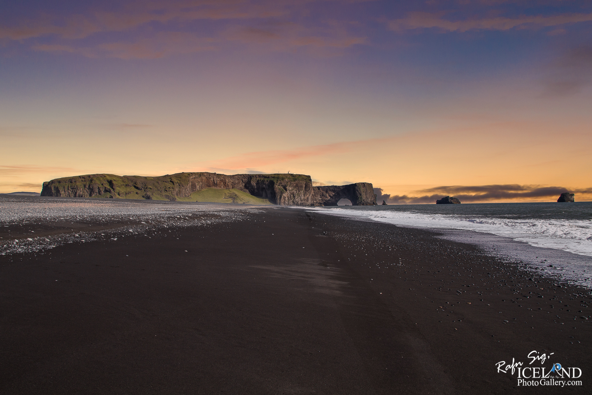 A dramatic coastal scene featuring a wide expanse of black sand beach. In the distance, a rugged cliff rises, partly covered in greenery, with a large rock formation that has a natural arch. The sky above displays a gradient of soft colors, transitioning from warm peach to cool blue as the sun sets. Gentle waves lap at the shore, and scattered pebbles dot the beach, enhancing the serene and untouched feeling of the landscape.
