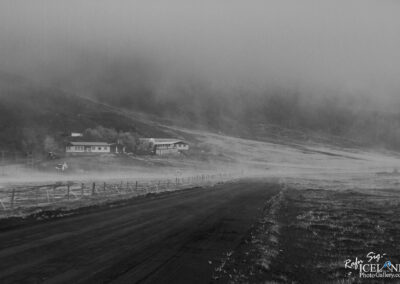 A winding dirt road stretches into the distance, bordered by a fence on one side. On the left, a cluster of simple buildings partially emerge from a low mist that blankets the landscape, giving the scene a mysterious atmosphere. The terrain slopes gently upward, leading to a fog-covered hill in the background. The image is black and white, enhancing the feeling of tranquility and isolation in this rural setting. The road appears slightly wet, hinting at recent rain, while the overall mood is quiet and serene, evoking a sense of calm and solitude in nature.