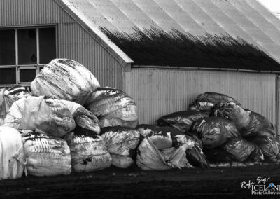 A large pile of assorted plastic bales, partially covered in dirt, sits against a corrugated metal building. The bales are wrapped in white and black plastic, showing signs of wear with stains and marks. Some bales are stacked high, creating an uneven, chaotic formation. In the background, a window with broken glass can be seen, and the roof of the building is angled, suggesting a rural or farm environment. The image is in black and white, emphasizing the textures and contrasts of the materials.