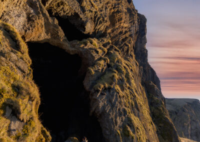 A rugged cliffside towers over a natural cave entrance. The cave's dark opening contrasts sharply with the warm colors of the grass and rocky surface. Sunlight casts soft shadows on the ground, revealing a path leading up to the cave. A solitary figure, likely a person, stands near the entrance, appearing small against the massive rock formation. The sky above showcases gentle hues of pink and blue, suggesting either dawn or dusk, adding a sense of tranquility to the scene.