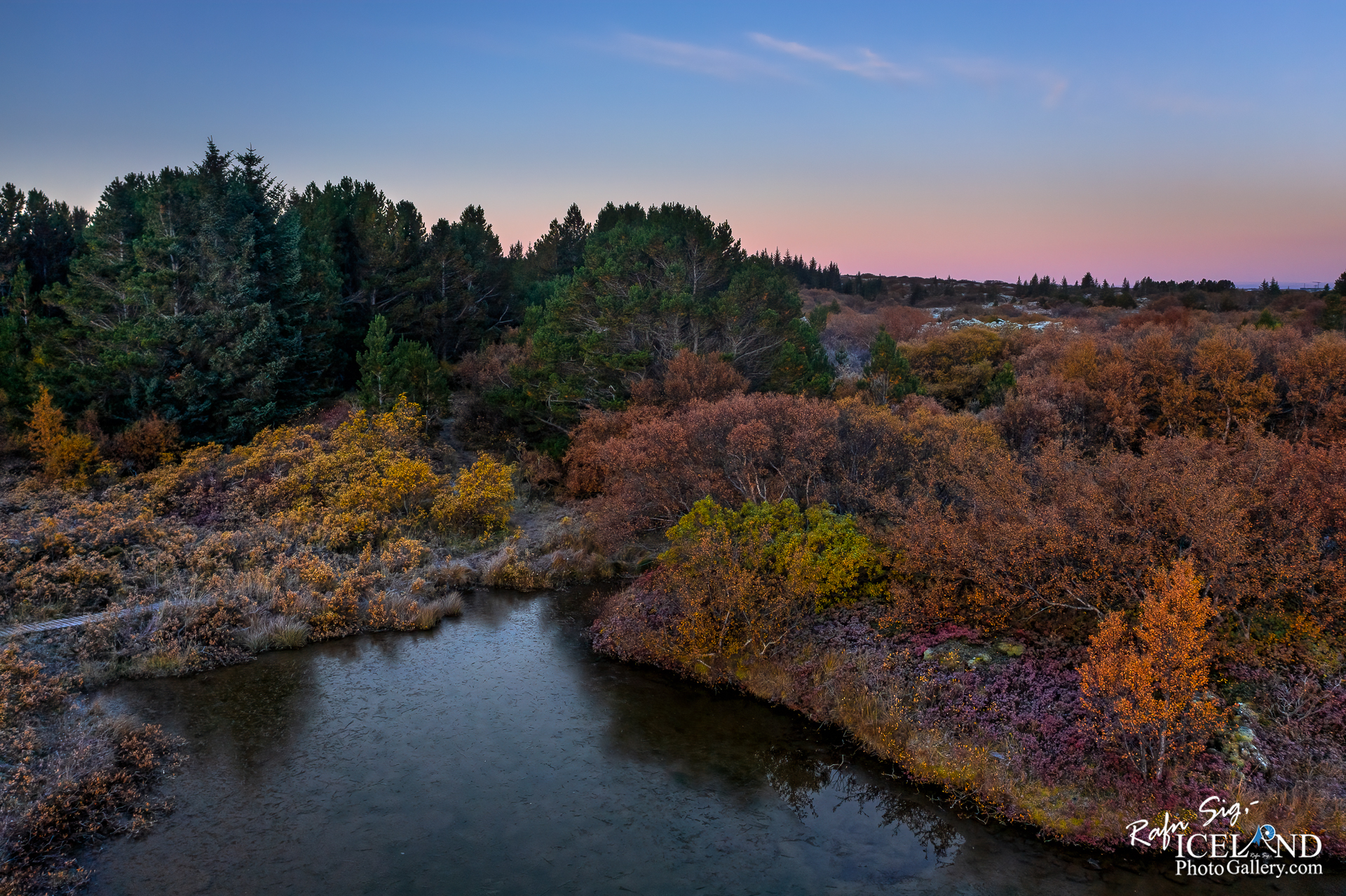 A serene lakeside scene featuring calm water that reflects the sky above, creating a mirror-like effect. The foreground has patches of grass and rocky terrain edging the water. In the background, gentle hills rise, partially covered in soft green vegetation and fading into the horizon. The sky is a mix of light blue and scattered clouds, suggesting a peaceful, early morning or late afternoon atmosphere. The overall mood conveys tranquility and natural beauty.