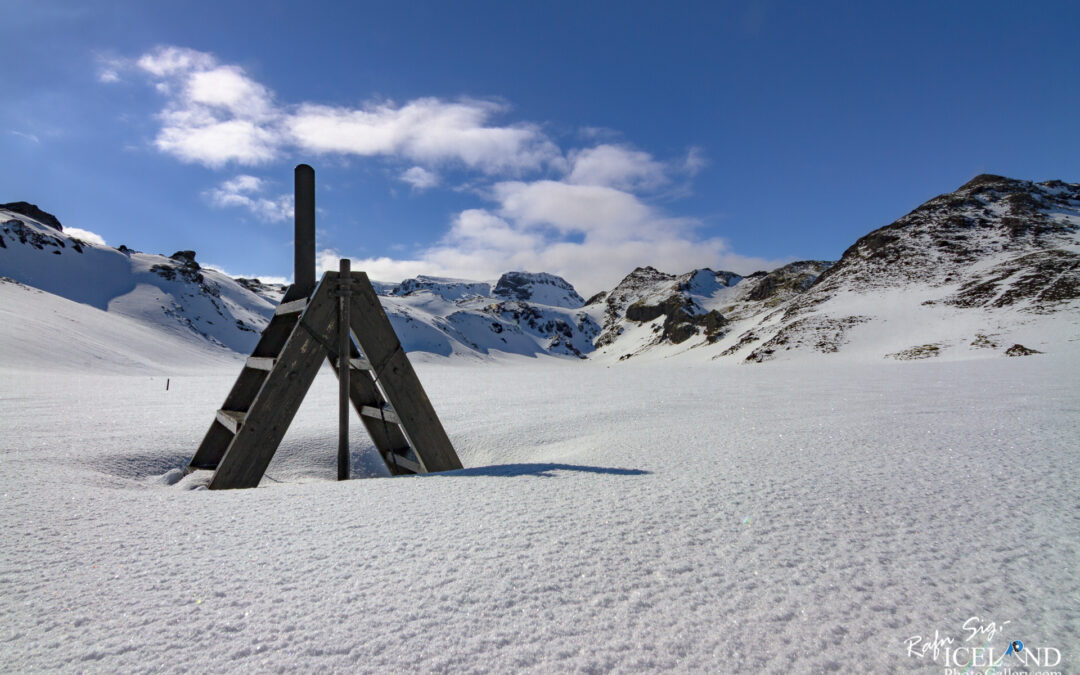 The image shows a snowy landscape under a bright blue sky with a few clouds scattered throughout. In the foreground, there is a wooden A-frame structure with a vertical pole at the top, standing on a blanket of pristine, white snow. The snow appears slightly sparkly, reflecting the sunlight. In the background, rugged snow-covered mountains rise, showcasing their peaks against the sky. The overall scene conveys a sense of tranquility and natural beauty in a chilly, winter environment.