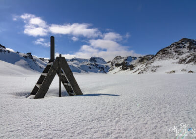 The image shows a snowy landscape under a bright blue sky with a few clouds scattered throughout. In the foreground, there is a wooden A-frame structure with a vertical pole at the top, standing on a blanket of pristine, white snow. The snow appears slightly sparkly, reflecting the sunlight. In the background, rugged snow-covered mountains rise, showcasing their peaks against the sky. The overall scene conveys a sense of tranquility and natural beauty in a chilly, winter environment.