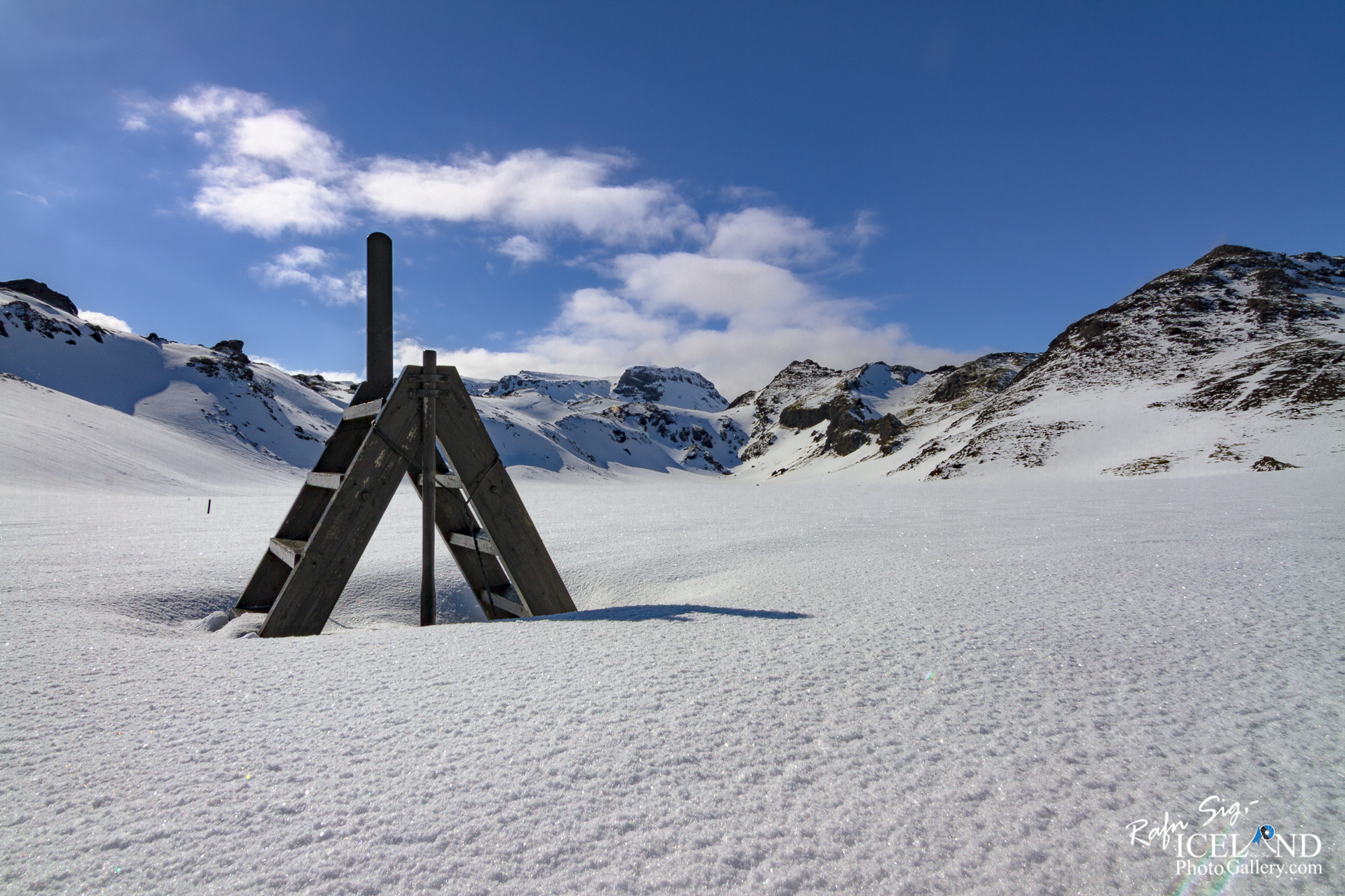 The image shows a snowy landscape under a bright blue sky with a few clouds scattered throughout. In the foreground, there is a wooden A-frame structure with a vertical pole at the top, standing on a blanket of pristine, white snow. The snow appears slightly sparkly, reflecting the sunlight. In the background, rugged snow-covered mountains rise, showcasing their peaks against the sky. The overall scene conveys a sense of tranquility and natural beauty in a chilly, winter environment.