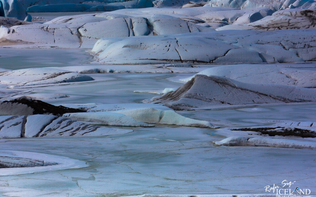 A vast, icy landscape featuring large, undulating mounds of snow and ice in various shades of white and pale blue. The surface is smooth and glossy in some areas, resembling frozen water, while other sections appear more rugged with gentle slopes and crevices. Small patches of darker earth peek through, adding contrast to the predominantly light colors. The sky above is mostly clear, enhancing the serene, untouched beauty of this wintery scene.