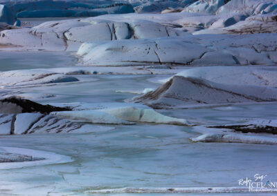 A vast, icy landscape featuring large, undulating mounds of snow and ice in various shades of white and pale blue. The surface is smooth and glossy in some areas, resembling frozen water, while other sections appear more rugged with gentle slopes and crevices. Small patches of darker earth peek through, adding contrast to the predominantly light colors. The sky above is mostly clear, enhancing the serene, untouched beauty of this wintery scene.
