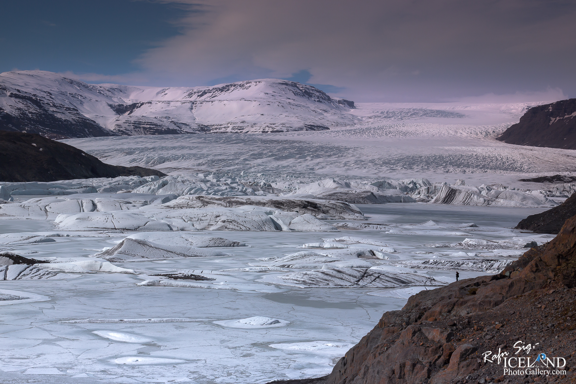 A vast, icy landscape featuring a massive glacier stretches across the scene, with layers of ice forming intricate patterns on its surface. The glacier appears to merge into a frozen lake below, surrounded by dark, rocky terrain. In the background, towering snow-capped mountains rise, their peaks glistening under a soft, cloudy sky. The overall color palette consists of vibrant whites, soft blues, and earthy tones, creating a stunning contrast between the natural elements. A small silhouette of a person stands on the rocky edge, emphasizing the scale of the expansive wilderness.