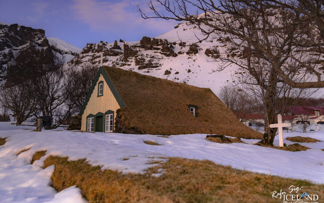 A small Church house with a thatched roof covered in grass sits nestled against a snow-covered landscape. The house features warm-colored wooden siding and has multiple windows framed in green. In the foreground, patches of snow contrast with the grassy areas, adding texture to the scene. On the left side, a bare tree’s branches stretch out, while a simple white cross stands nearby, suggesting a tranquil setting. In the background, rugged mountains rise, partially covered in snow, under a pink-hued sky. The overall ambiance is serene and picturesque, evoking a sense of peaceful isolation in nature.