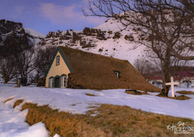 A small Church house with a thatched roof covered in grass sits nestled against a snow-covered landscape. The house features warm-colored wooden siding and has multiple windows framed in green. In the foreground, patches of snow contrast with the grassy areas, adding texture to the scene. On the left side, a bare tree’s branches stretch out, while a simple white cross stands nearby, suggesting a tranquil setting. In the background, rugged mountains rise, partially covered in snow, under a pink-hued sky. The overall ambiance is serene and picturesque, evoking a sense of peaceful isolation in nature.