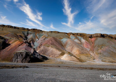 A dramatic landscape features a series of colorful, layered hills with rich hues of red, orange, green, and beige, resembling a natural canvas. The hills slope downward, revealing textures and folds in the rock. Above, a bright blue sky is adorned with wispy white clouds, creating a striking contrast against the earthy tones of the terrain below. In the foreground, a rocky surface is scattered with smaller stones, enhancing the natural beauty of this rugged environment. The overall scene conveys a sense of vastness and tranquility in nature.