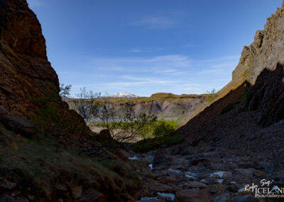 A narrow rocky pathway leads through steep, rugged cliffs, revealing a vast landscape beyond. The foreground features a stony riverbed with scattered rocks and patches of greenery, including small bushes. In the background, colorful hills adorned with shades of purple, green, and brown rise majestically, with a snow-capped peak visible in the distance under a clear blue sky. The scene conveys a sense of natural beauty and tranquility.