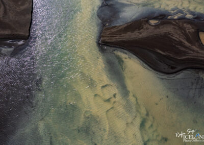 An aerial view of a river delta showcases a striking contrast between water and land. The water is a blend of colors, ranging from deep teal to lighter greens and browns, creating a shimmering effect as sunlight reflects off its surface. Geometric patterns emerge as the river meanders, flowing between dark, sandy banks made of textured earth. Sections of the delta appear smooth and reflective, while others exhibit gentle ripples, adding depth and dimension to the landscape. The interplay of color and form presents a captivating scene of natural beauty.