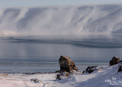 A serene, icy landscape unfolds, featuring a frozen lake surrounded by snow-covered hills and rocky formations. In the foreground, a prominent rock stands out, partially dusted with snow, while the background displays smooth, rolling hills that blend into the pale blue sky. The surface of the lake reveals intricate patterns created by ice, reflecting the calm ambiance of the winter scene. The overall atmosphere is tranquil, evoking a sense of nature's stillness in a cold, remote area.