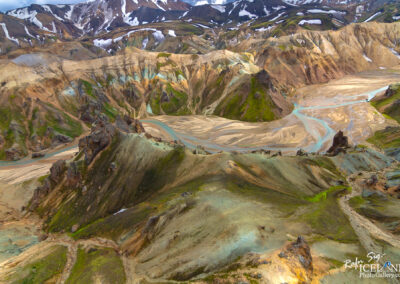 A panoramic view of a rugged mountainous landscape featuring a variety of colors. The terrain is characterized by undulating hills, some covered in smooth, green vegetation, while others are stark and rocky in shades of brown, gold, and grey. The ground is marked by curving patterns, suggesting paths or waterways that wander through the scene. Snow patches linger in the higher elevations, contrasting against the earthy tones of the mountains. The sky above is partially cloudy, casting dynamic shadows across the landscape, enhancing the dramatic feel of this natural setting.