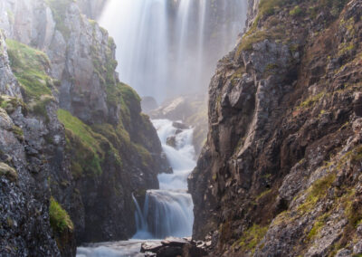 A stunning landscape featuring a cascading waterfall surrounded by rugged cliffs. The water flows down several tiers, creating a series of smaller falls and gentle rapids. The rocks are dark and textured, partially covered with vibrant green moss. In the background, soft green hills rise, creating a serene and inviting atmosphere. The scene is illuminated by a bright light, suggesting early morning or late afternoon, giving the water a shimmering quality. Mist rises from the falls, adding a sense of movement and freshness to the air.