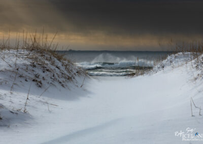 A snowy landscape leads to the ocean, framed by patches of tall grass on either side. The beach is covered in thick, white snow, creating a smooth and serene pathway toward the water. Gentle waves can be seen rolling in, their surfaces glistening subtly under a mix of gray and soft orange clouds above. The atmosphere feels calm yet dynamic, as the waves break softly, contrasting with the stillness of the snow-covered ground.