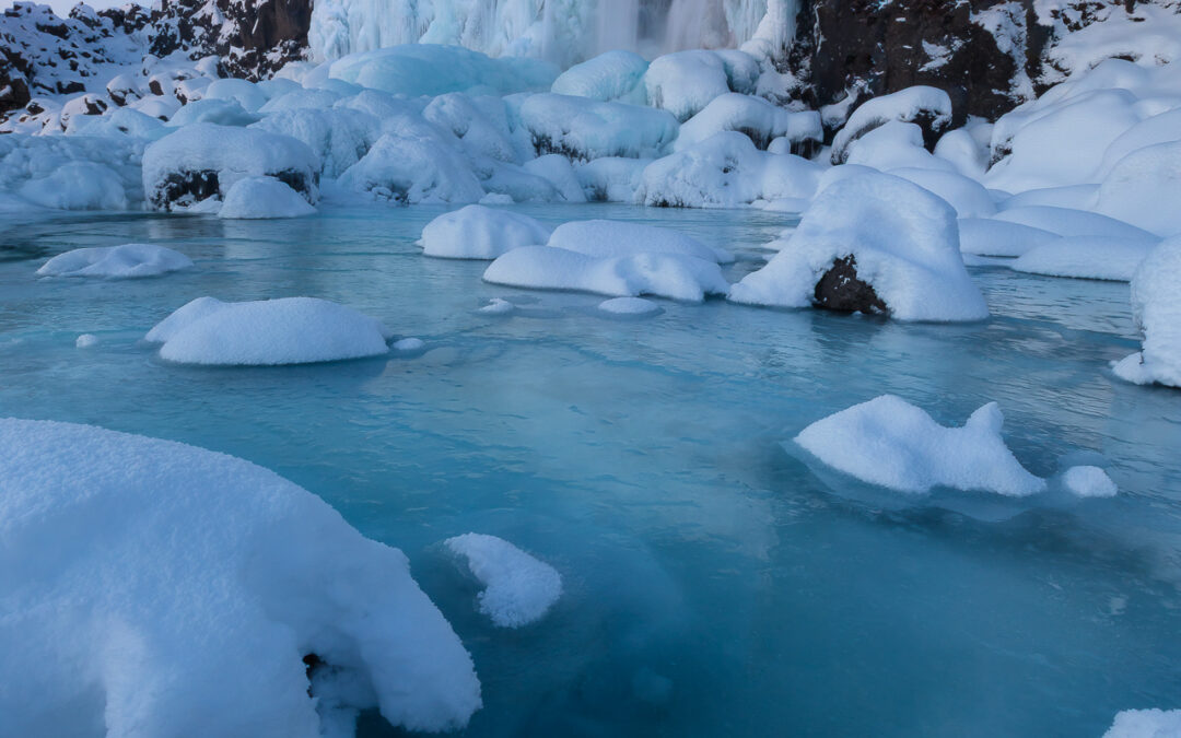 A serene winter landscape features a majestic waterfall cascading down a rocky cliff, surrounded by ice and snow. The water appears to glisten as it flows over the frozen edges, creating a stunning visual contrast against the dark rocks. Small ice formations dot the foreground, with soft, fluffy snow covering them. A still, icy blue pool of water reflects the pale sky above, enhancing the tranquil and frosty atmosphere of this wintry scene.