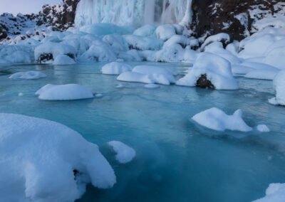 A serene winter landscape features a majestic waterfall cascading down a rocky cliff, surrounded by ice and snow. The water appears to glisten as it flows over the frozen edges, creating a stunning visual contrast against the dark rocks. Small ice formations dot the foreground, with soft, fluffy snow covering them. A still, icy blue pool of water reflects the pale sky above, enhancing the tranquil and frosty atmosphere of this wintry scene.