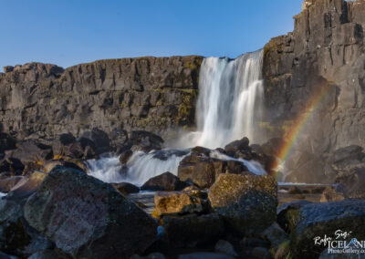 A majestic waterfall cascades down from a rocky cliff, surrounded by dark, uneven rocks and patches of green moss. The water flows vigorously, creating a misty spray that catches the sunlight, producing a faint rainbow at its base. The sky above is clear and blue, contrasting sharply with the rugged greys and browns of the rocks. The scene captures the serene power of nature, with the sound of rushing water adding to the tranquil ambiance.