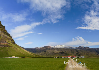 A panoramic view of a dramatic landscape featuring a prominent, steep, green mountain rising sharply from a flat, expansive grassland. The mountain, with rocky textures and a gentle cover of greenery, stands against a backdrop of blue sky adorned with scattered white clouds. In the foreground, a pathway leads towards a small cluster of buildings nestled at the base of the mountain, surrounded by vibrant green fields. The scene evokes a sense of tranquility and the grandeur of nature, highlighting the contrast between the rugged terrain and the smooth, open fields.