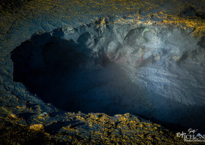 The image depicts a close-up view of a geothermal hot spring crater. The ground surrounding the crater is cracked and uneven, showing a mix of textured earth tones, including dark blues and warm golden hues. The crater itself is somewhat circular and appears deep, with a light mist rising from the water inside, suggesting heat. The overall scene conveys an otherworldly, natural landscape, with an emphasis on the contrast between the dry ground and the wet, steamy opening of the spring.