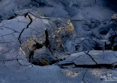 The image showcases a rugged geothermal landscape characterized by cracked, dark grayish-black earth showing signs of heat and steam activity. The surface is fissured with deep crevices, and some areas appear to have a slightly shiny texture, reflecting the moisture beneath. Pockets of bubbling mud and shallow pools can be seen, hinting at the geothermal activity in the area. Along the edges, there are areas where the ground is uneven, creating a rugged and raw appearance, while some spots display hints of yellowish mineral deposits. The overall atmosphere conveys a sense of otherworldliness, typical of volcanic terrains.
