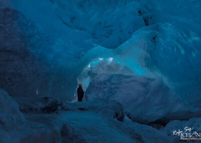 The image depicts a stunning ice cave illuminated by a soft blue light. Tall, towering ice formations create a cavernous space with smooth, glacial surfaces. In the foreground, a person wearing a dark jacket and a helmet stands, gazing at the icy landscape around them. The scene feels both expansive and intimate, with the blue hues evoking a tranquil yet majestic atmosphere. The intricate textures of the ice add depth, while the light highlights the cave’s natural beauty and allure.