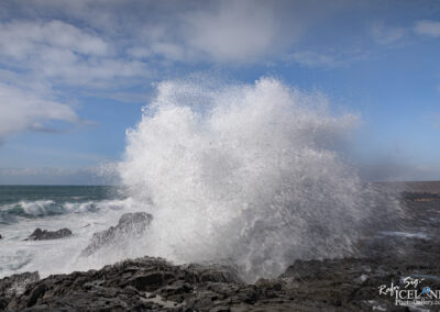 A powerful ocean wave crashes against dark, rugged rocks, sending a spray of white water high into the air. In the background, the ocean stretches out, with rolling waves and a bright blue sky dotted with soft clouds. The scene captures the dynamic energy of the sea, showcasing the contrast between the dark shoreline and the vibrant water and sky.