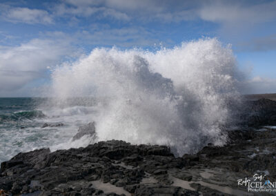 The image captures a dramatic seascape where powerful ocean waves crash against rugged black rocks. Water is propelled high into the air, creating a misty spray that glimmers in the sunlight. The background features a vast expanse of turquoise water under a partly cloudy sky, adding depth to the scene. The contrast between the dark, jagged rock formations and the bright white foam of the waves evokes a sense of raw energy and natural beauty.