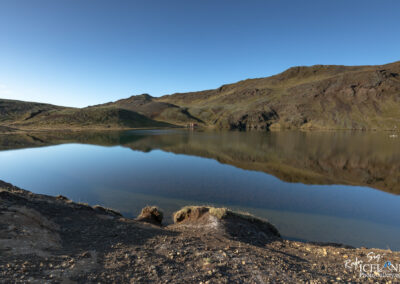 A serene landscape features a calm lake surrounded by rolling hills and rocky terrain. The water reflects the blue sky above, creating a mirror-like effect. On the far side, a small rustic cabin can be seen quietly nestled among the hills, adding a touch of charm to the scene. The hills vary in color, showcasing greens and browns, while gentle slopes meet the tranquil waters, enhancing the peaceful atmosphere of this natural setting. The scene conveys a sense of stillness and beauty.
