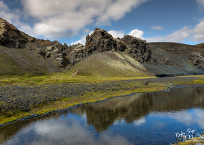 A serene landscape featuring rugged, rocky hills and a calm body of water in the foreground. The hills are varied in color, with dark volcanic rock and patches of vibrant green moss. The sky above is partly cloudy, with soft clouds casting shadows over the terrain. The water reflects the surrounding environment, creating a peaceful, mirrored effect. The scene conveys a sense of tranquility and the raw beauty of nature.